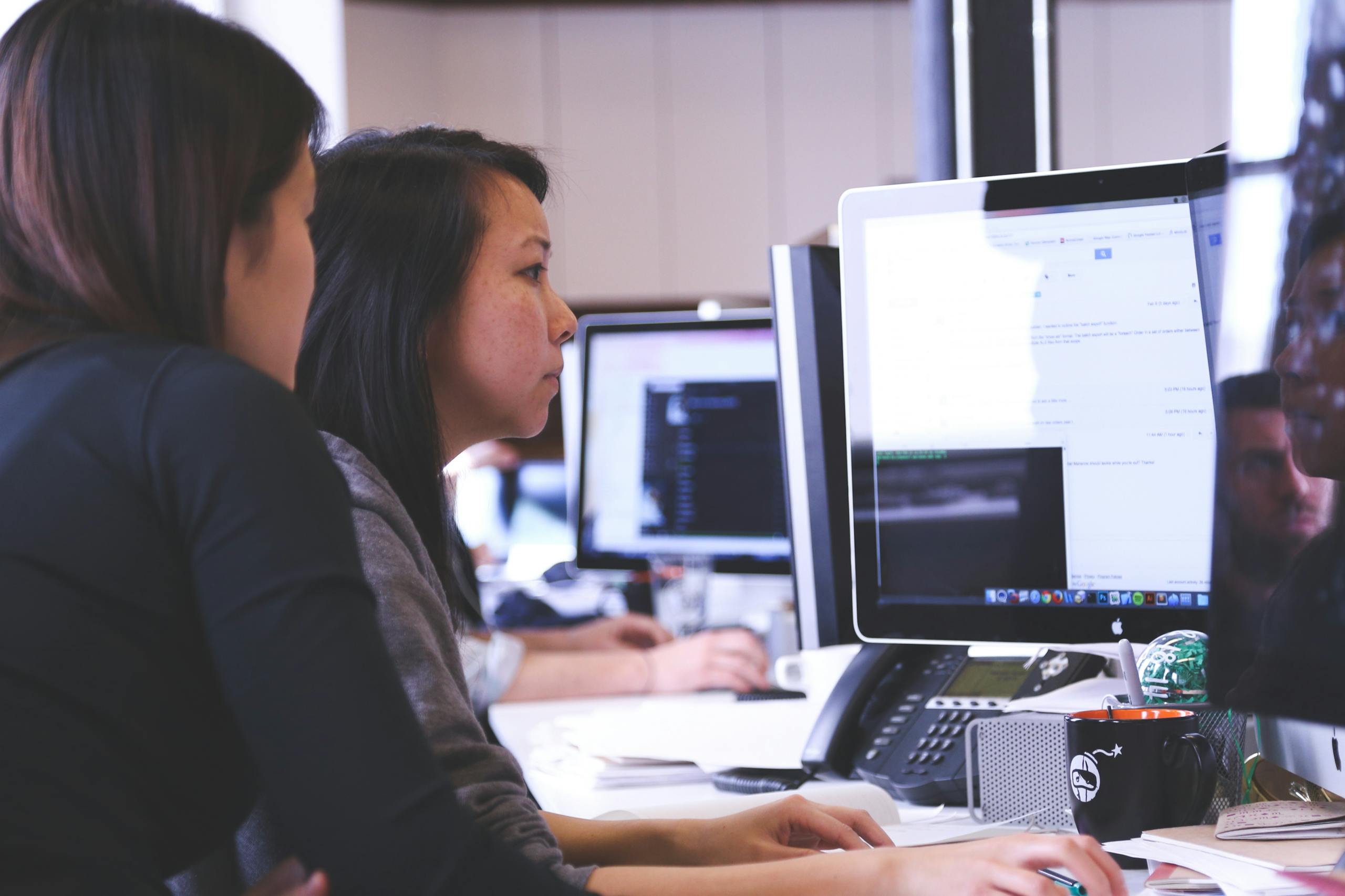 Two women working together on code at a computer in a modern office setting.
